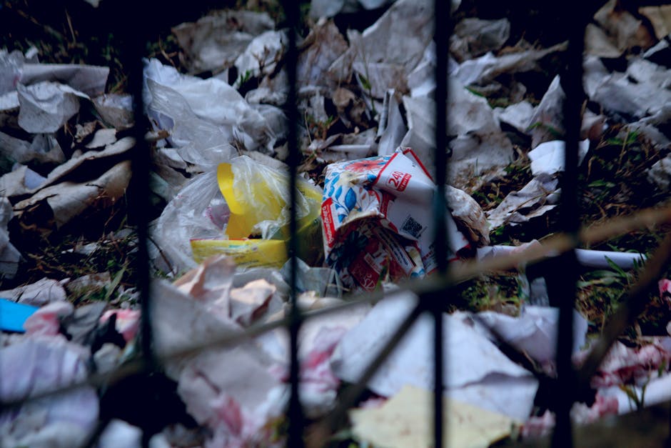 A person's hand, wearing a beige rubber glove, holds a yellow waste collection container labeled 'WASTE' in a laboratory or clinical setting. The container is filled with used pipettes and other small