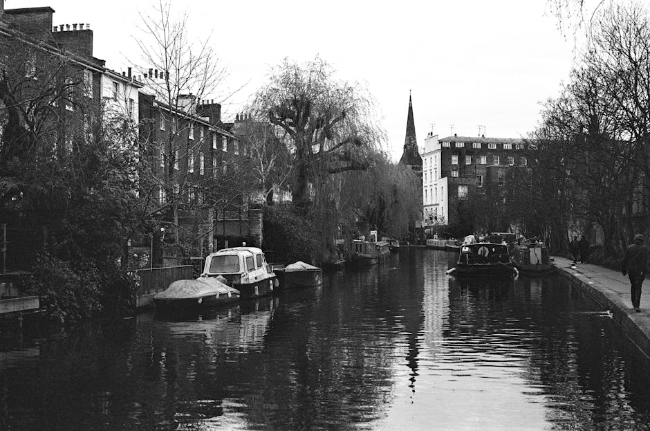 A black and white photograph of a narrow canal in Little Venice, Paddington, with several small boats moored along the water's edge. The canal is bordered by residential buildings with brick facades on the left side and a paved walkway on the right, where a few pedestrians are visible walking. Tall, leafless trees line both sides of the canal, and a church steeple can be seen in the background, rising above the surrounding structures. The water appears calm with gentle ripples, reflecting the surrounding trees and buildings, and the scene is illuminated by natural daylight, creating a serene, quiet atmosphere. Although the image does not depict cleaning directly, it highlights the importance of maintaining cleanliness and hygiene in residential exterior environments, aligning with the expertise of Paddington Cleaners in surface cleaning and property maintenance, especially relevant for homes in picturesque areas like Little Venice.