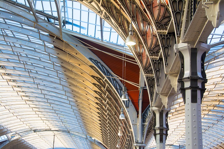 The image depicts an indoor train station platform with a high, arched glass and metal roof allowing natural light to illuminate the area. The platform surface is made of polished tiles, reflecting th