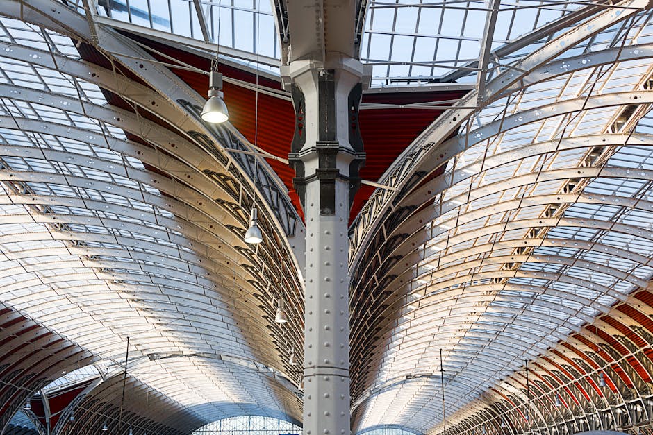 Interior view of a large, modern architectural structure featuring a high, curved ceiling with a series of steel beams and glass panels. The ceiling is illuminated by hanging industrial-style pendant 